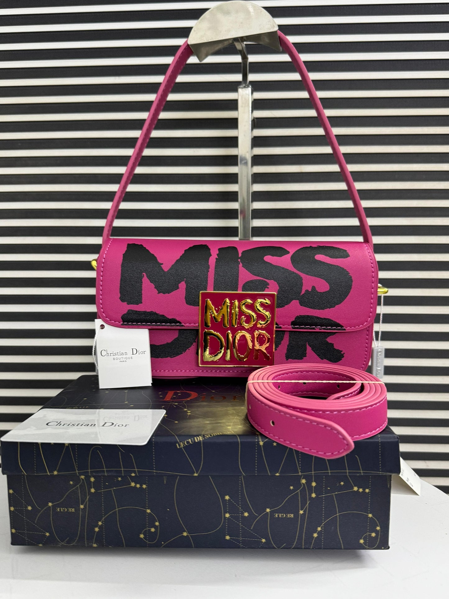 Pink handbag with 'MI SS DI OR' text, belt, and Dior box on a striped background