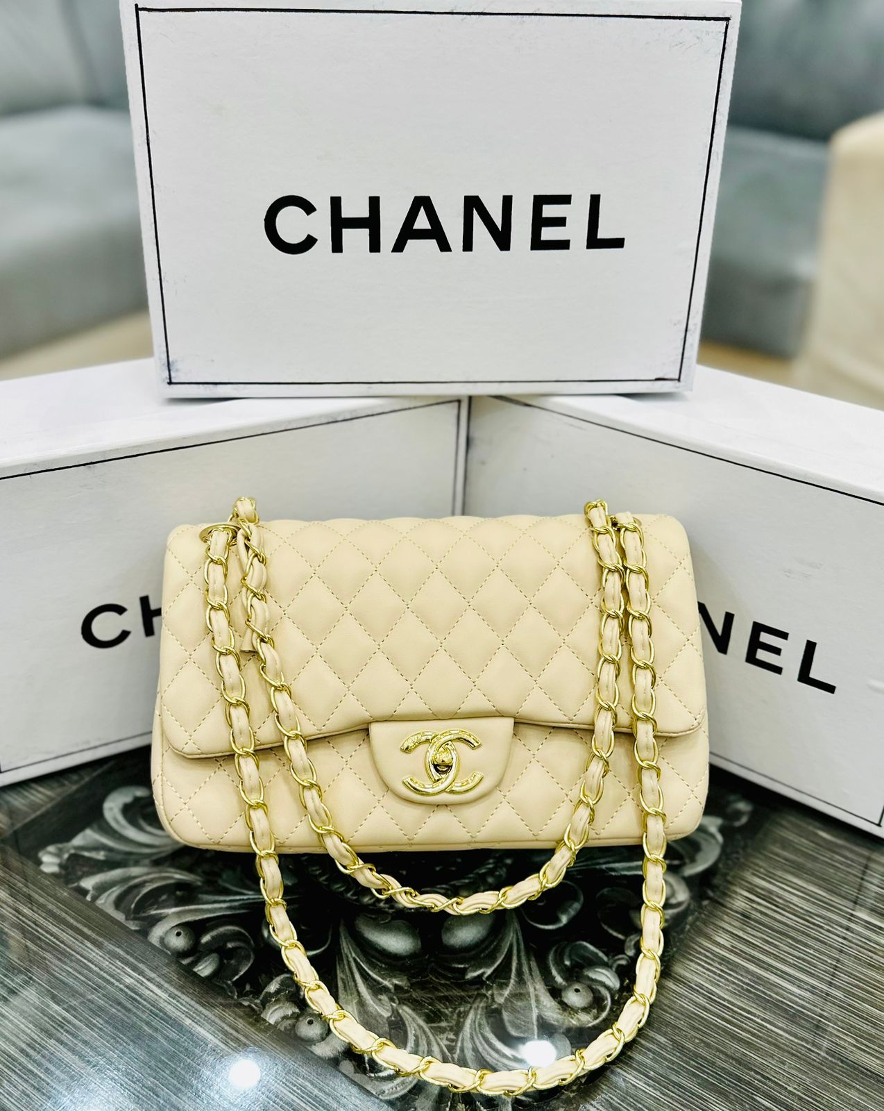 Beige Cha nel handbag with gold chain on a reflective surface with 'Cha nel' branding in the background.