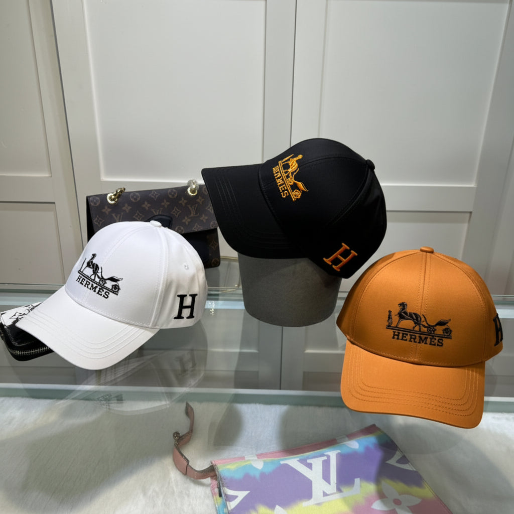 Four embroidered baseball caps displayed together on a glass surface in front of a white paneled wall. The caps are white, black, beige, and orange, each featuring an embroidered Hermes logo and horse-and-carriage emblem on the front and side.
