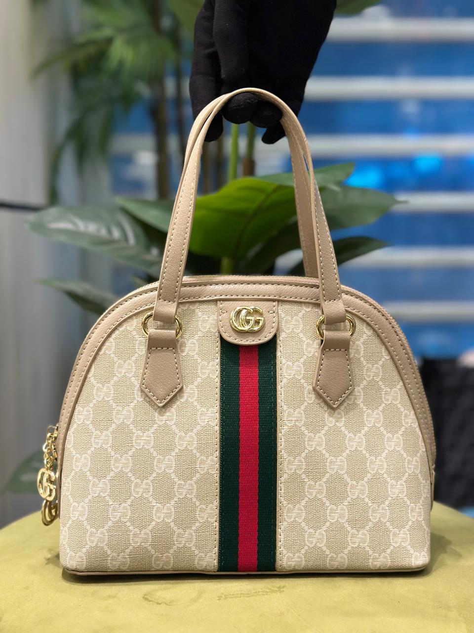 Beige handbag with a red and green stripe held by a gloved hand against a blurred indoor background.