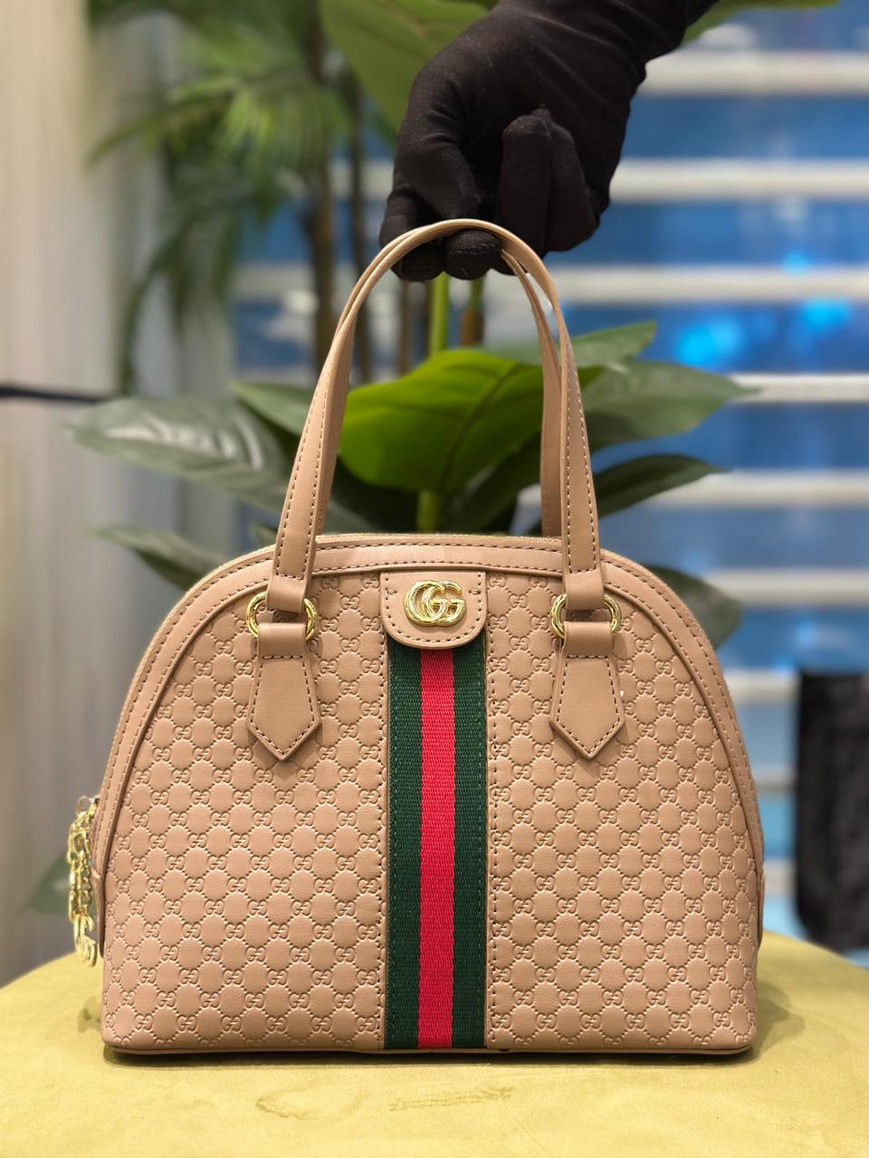 Beige handbag with a red and green stripe held by a gloved hand against a blurred indoor background