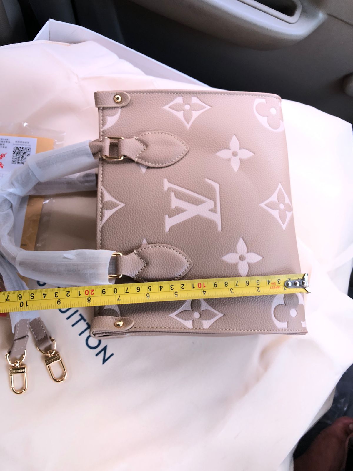 Beige handbag with gold hardware on a white surface, measured by a yellow tape measure.
