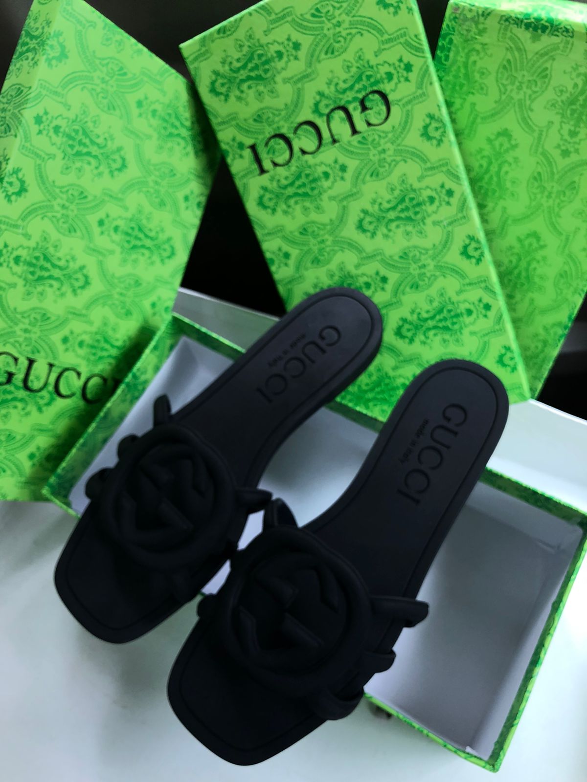 Black sandals with floral details on a white surface, with Guc ci green boxes in the background.