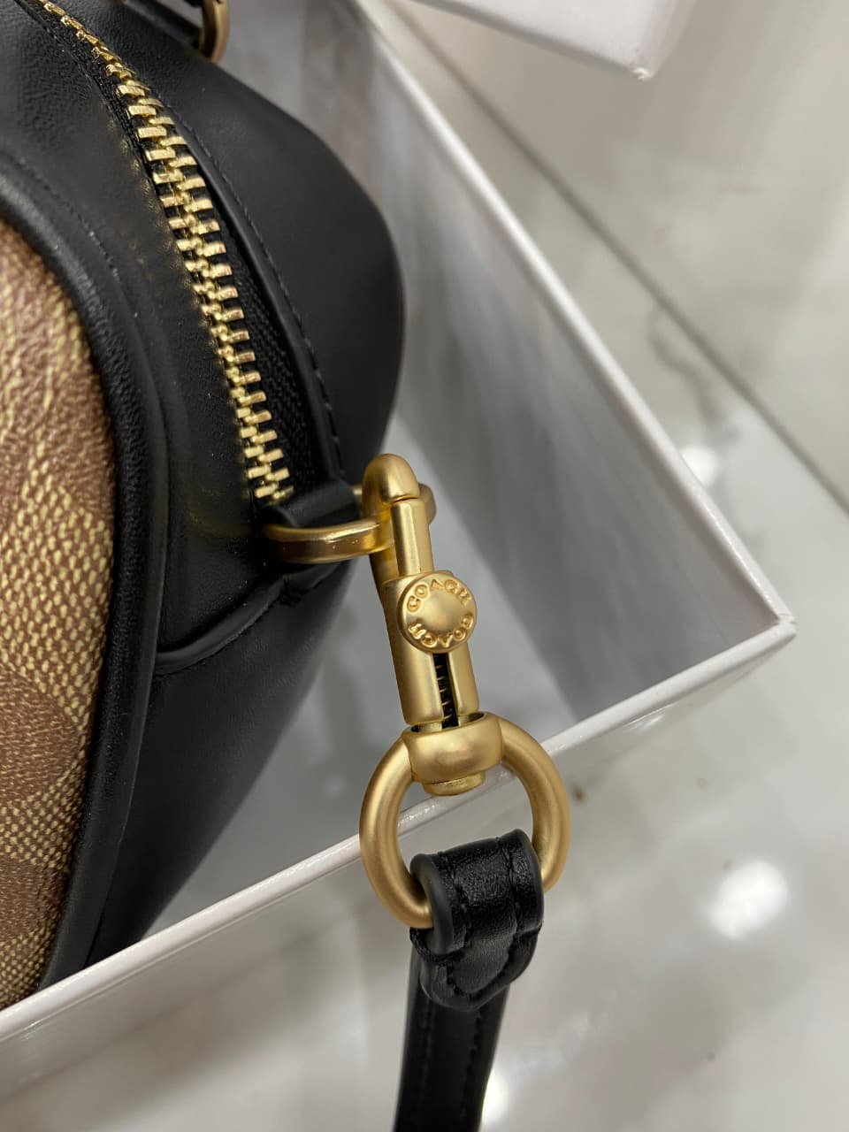 Close-up of a black handbag with gold zipper pull and strap on a reflective surface.