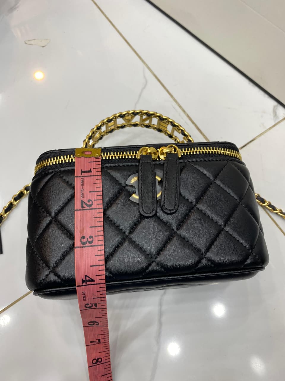Black quilted handbag with gold chain and zipper, measured with a red tape measure on a white background.