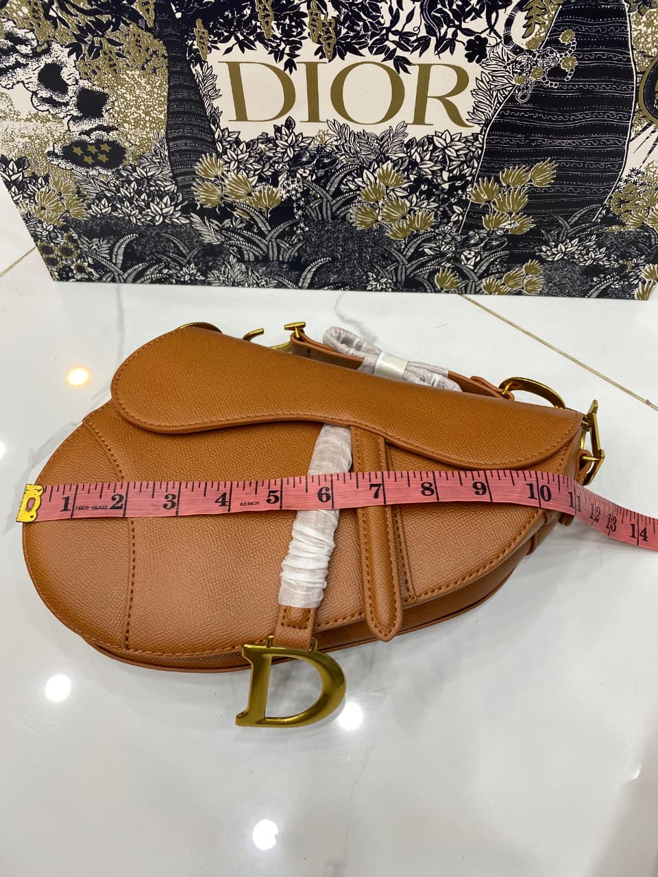 Brown leather bag with Di or logo and measuring tape, against a floral Di or box background.