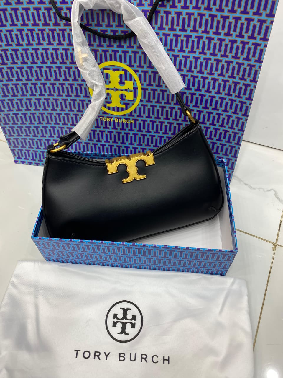Black handbag with gold logo on a To ry Bur ch branded box and white cloth.