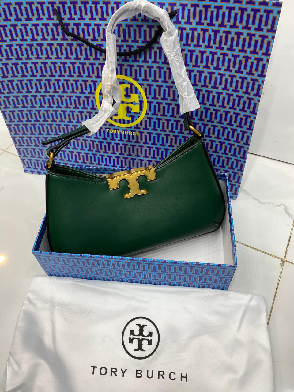 Green handbag with gold clasp on a white surface, with a blue To ry Bur ch branded box in the background.