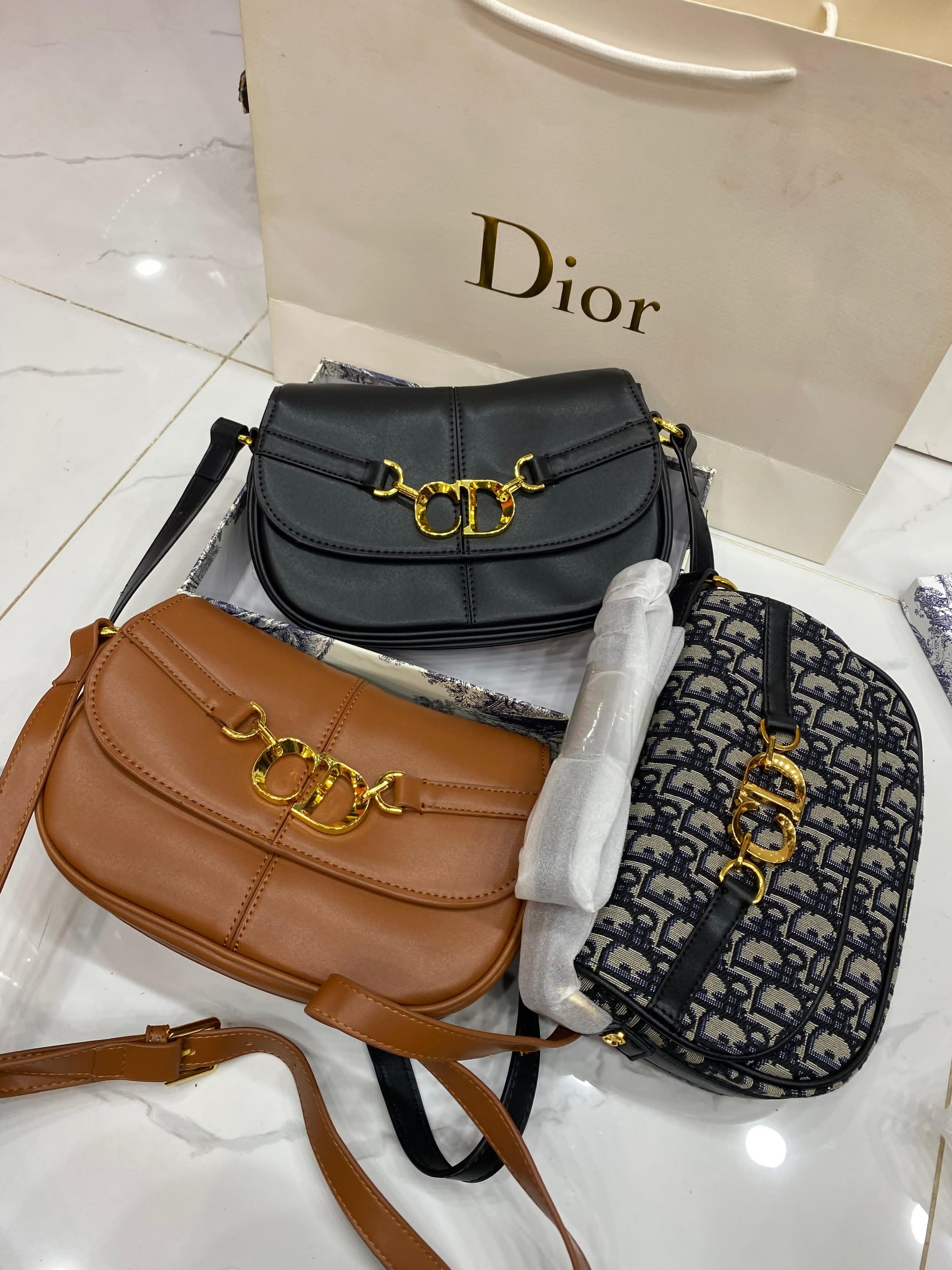 Two handbags, one brown and one black with gold hardware, on a white surface with a Dior shopping bag in the background.