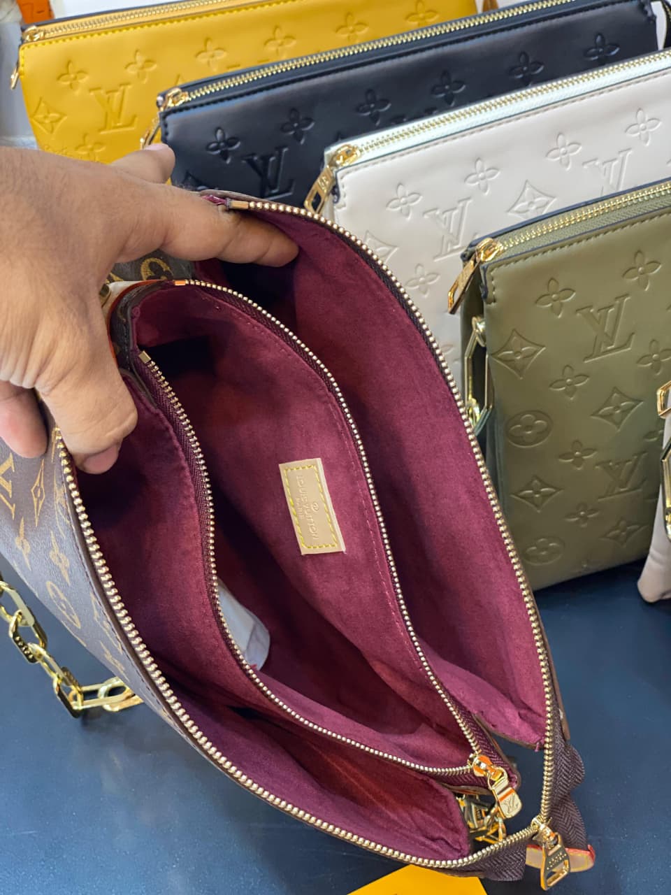 Close-up of a hand holding an open maroon handbag with visible brand logo, set against a background of other handbags.