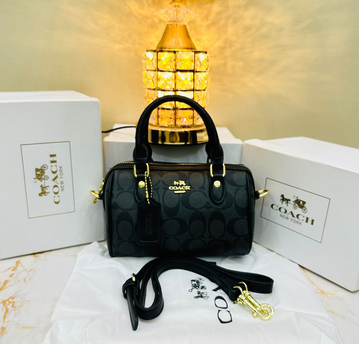 Black Co ach handbag with gold accents on a white surface with Co ach logo in the background.