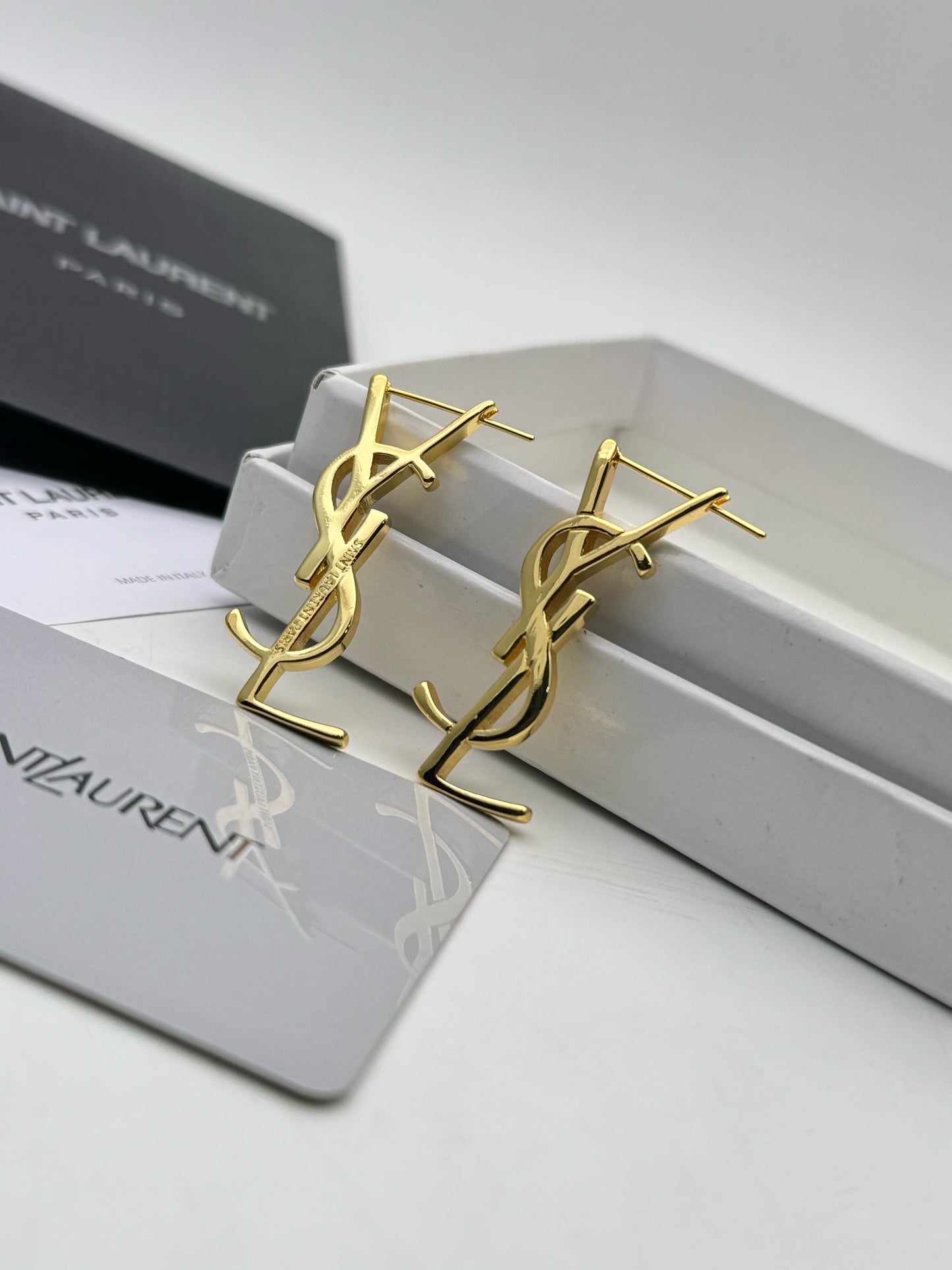 Gold Y S L earrings on a white surface with Yv es Sa int Lau rent branding.