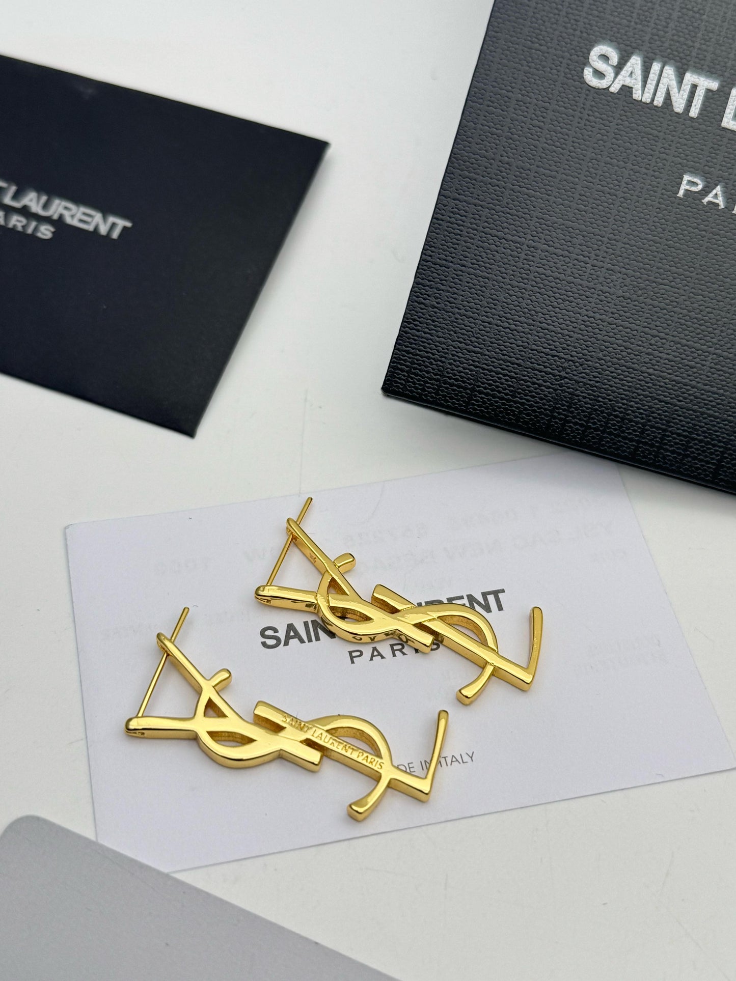Gold Y S L earrings on a Sa int Laur ent branded card with black boxes in the background.