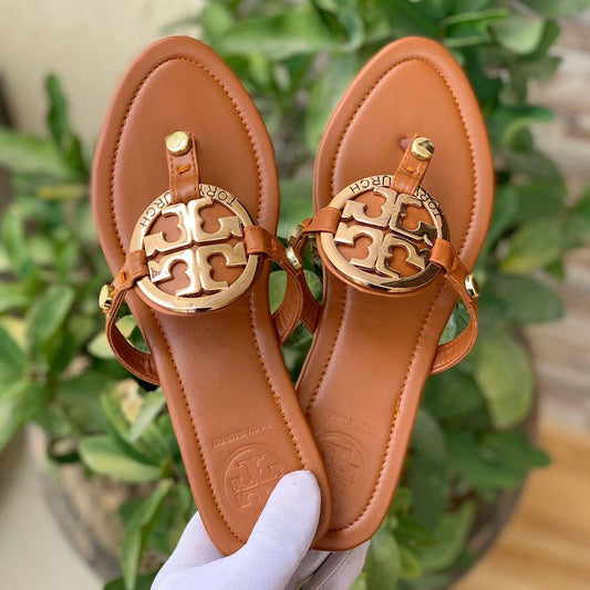 Pair of brown sandals with gold logo held by a hand against a green plant background