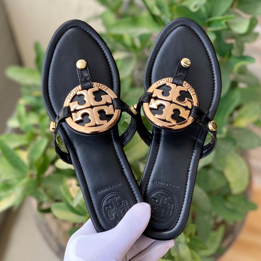 Black sandals with gold logo held by a hand against a green leafy background