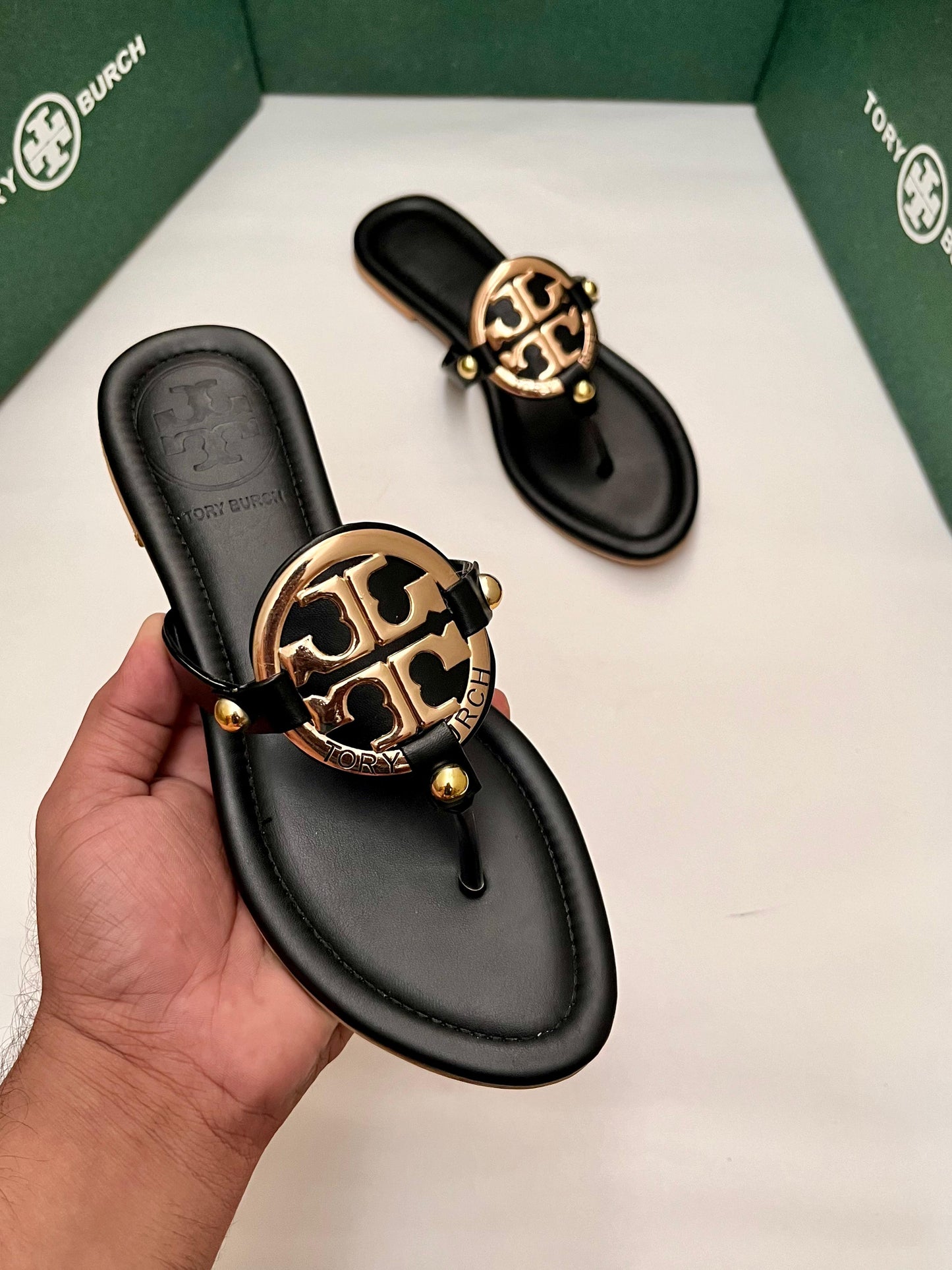 Black sandals with gold logo held by a hand, displayed against a white background.