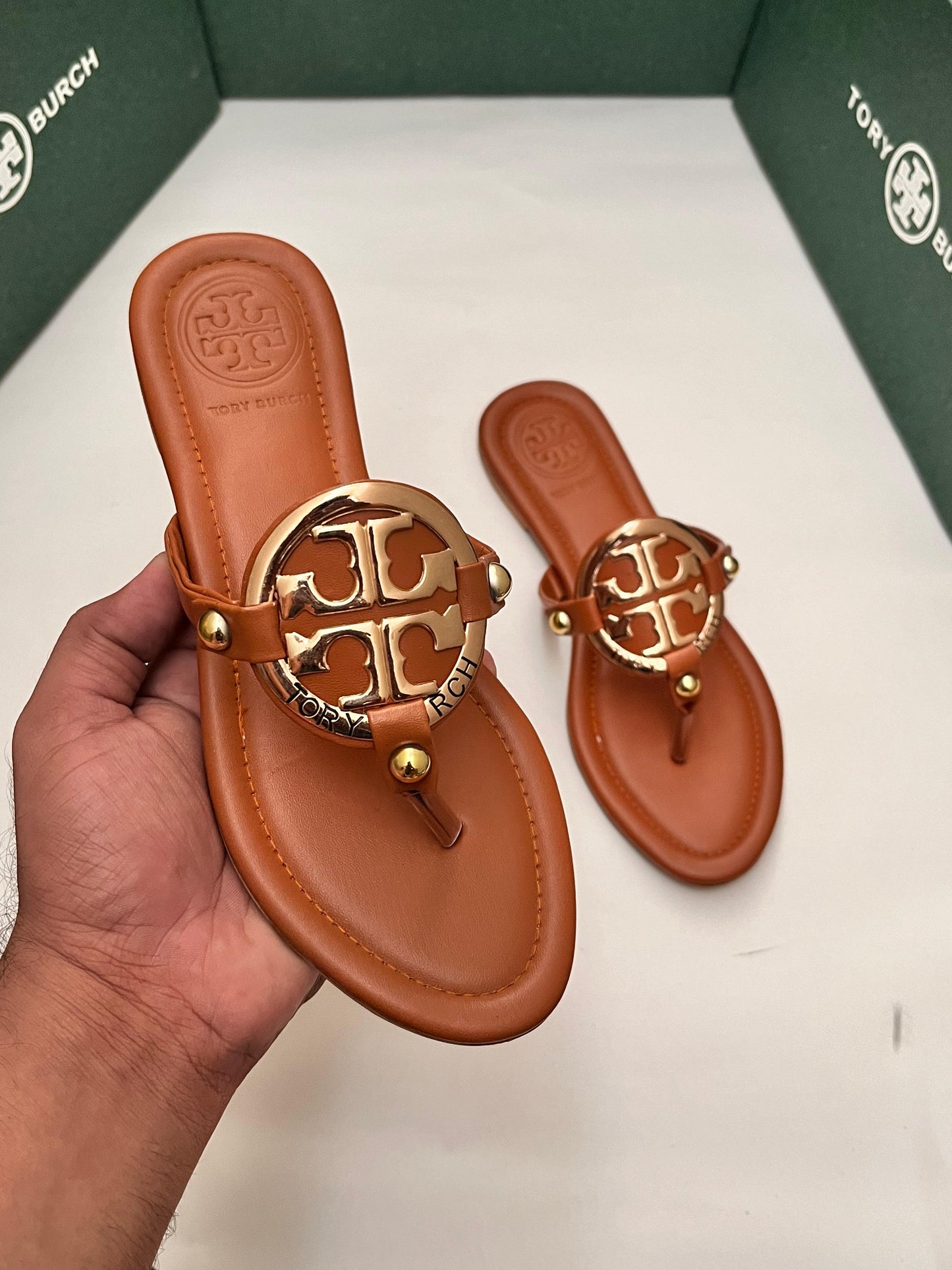 Brown sandal with gold logo held by a hand, brand box in the background