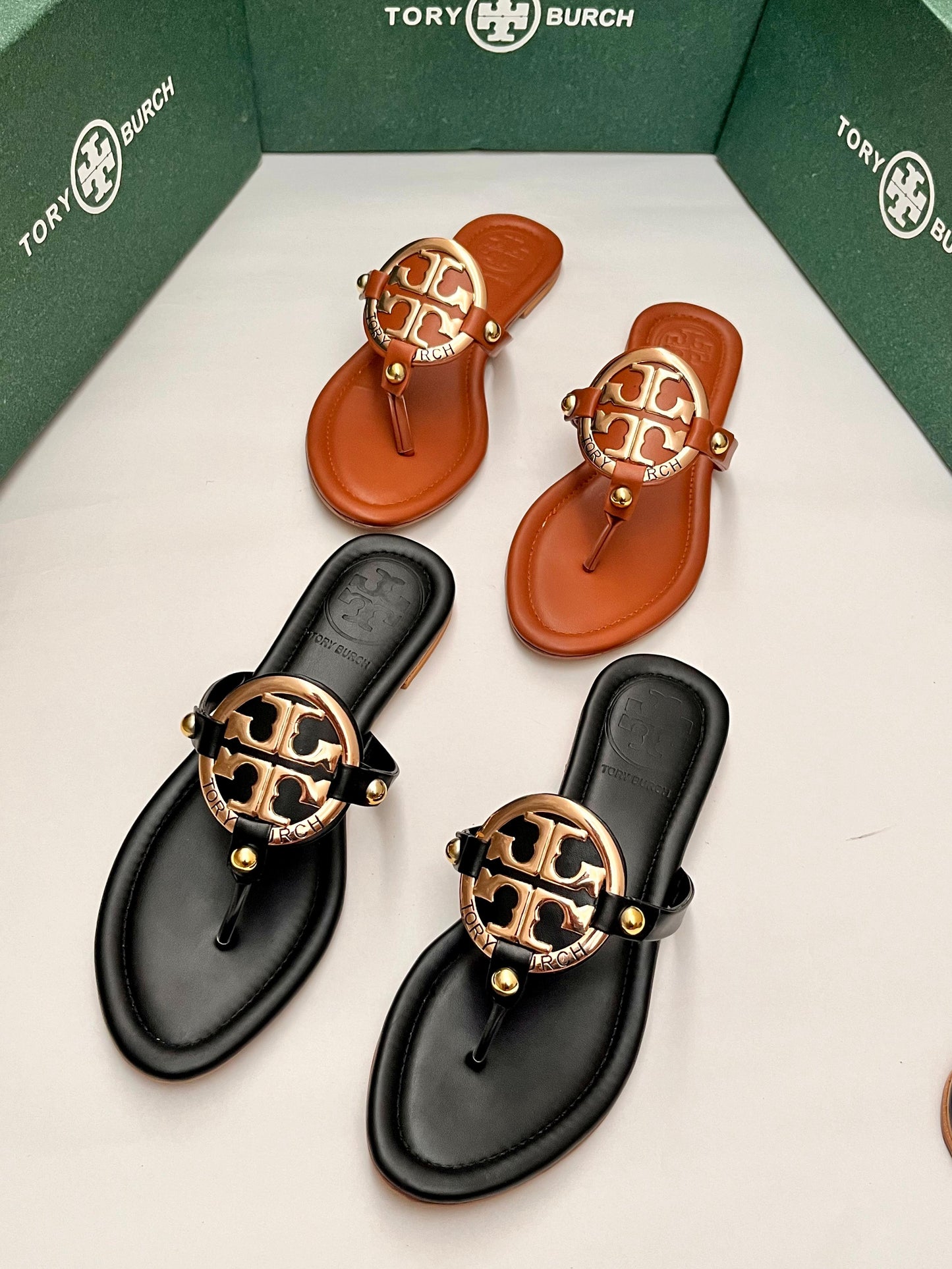 To-ry Bur-ch sandals in black, brown, and gold on a white surface with green box in the background.