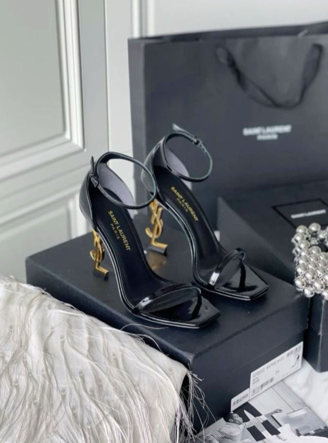 Black high-heeled sandals on a black box with Yv es Sa int Lau rent branding, next to a black handbag.