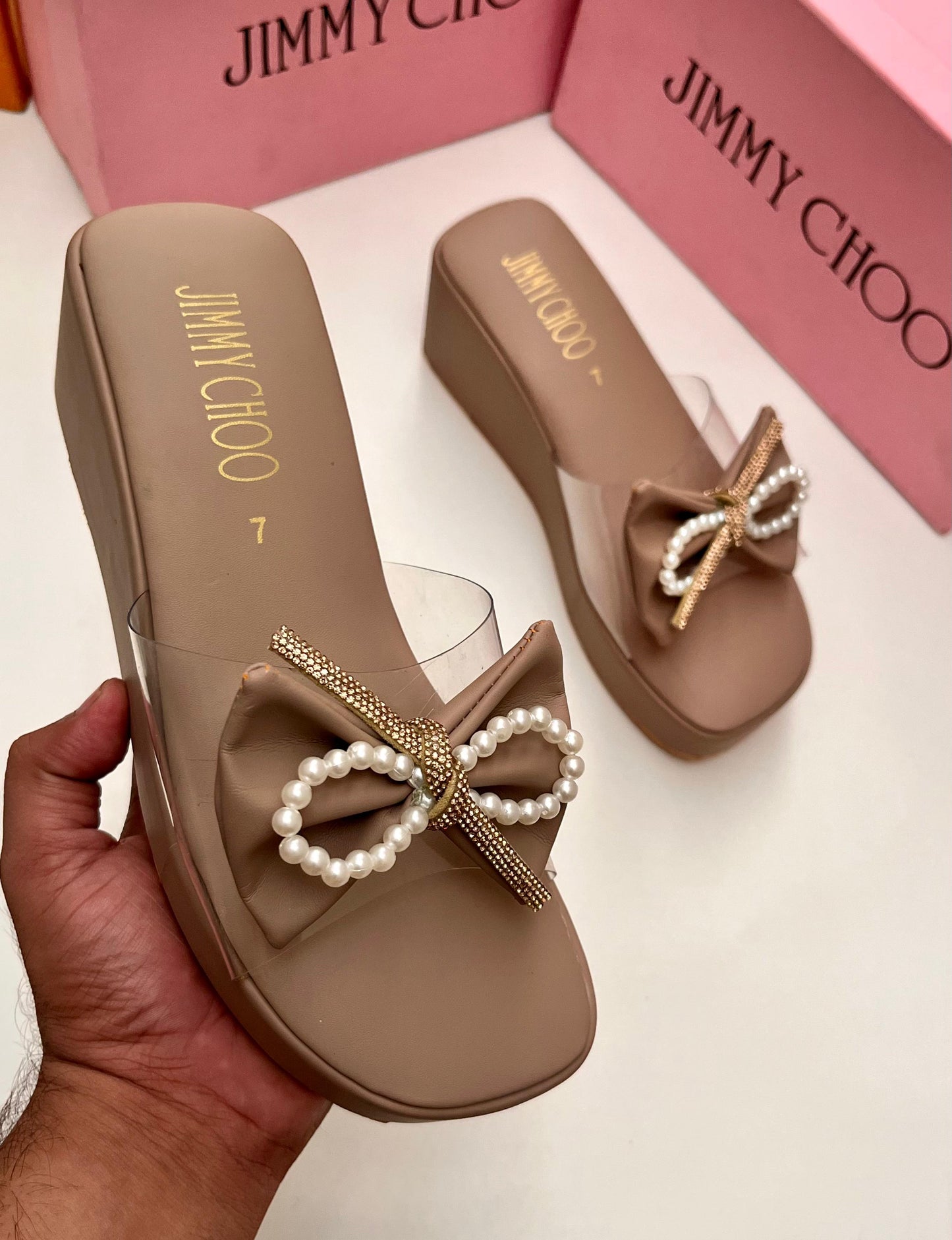 Beige sandals with decorative bows and pearls, held by a hand, with 'Jim my Ch oo' branding.