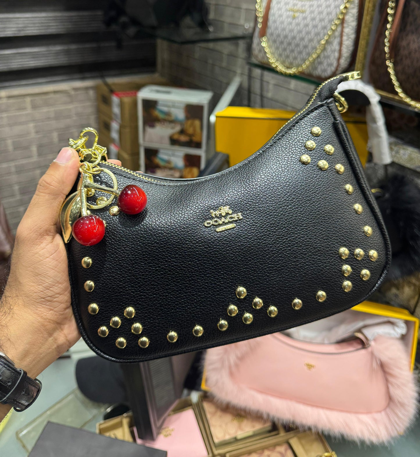 Black Co ach handbag with gold studs and red cherries held by a person in a store setting.