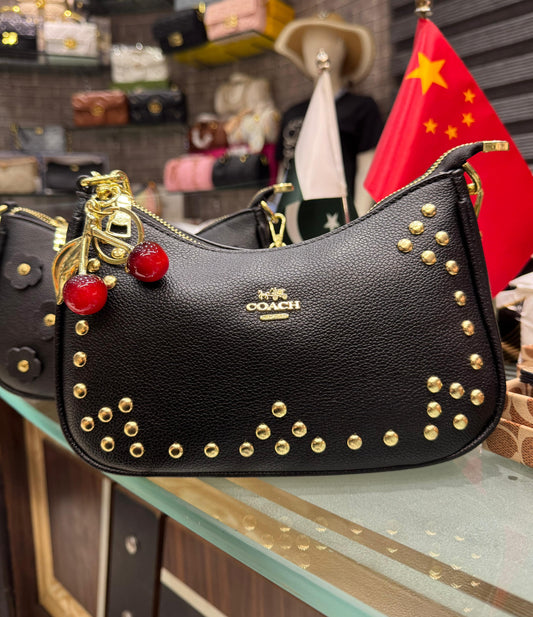 Black Coa ch handbag with gold studs and red accents on a glass surface, with a blurred background.