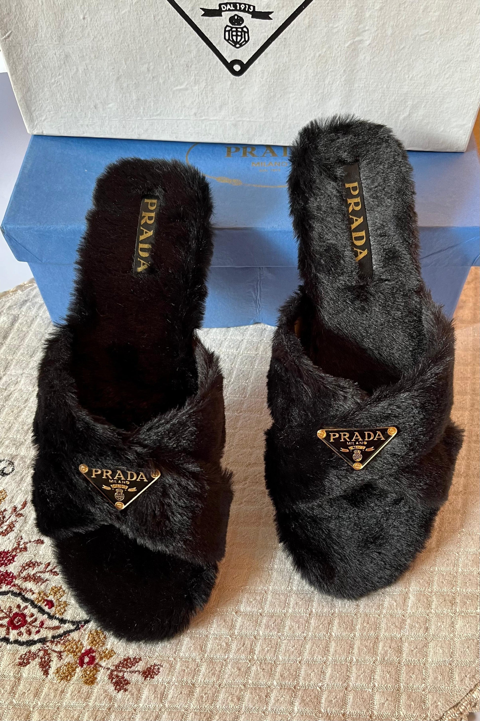 Black fuzzy Pra da slippers on a patterned surface with a box in the background.