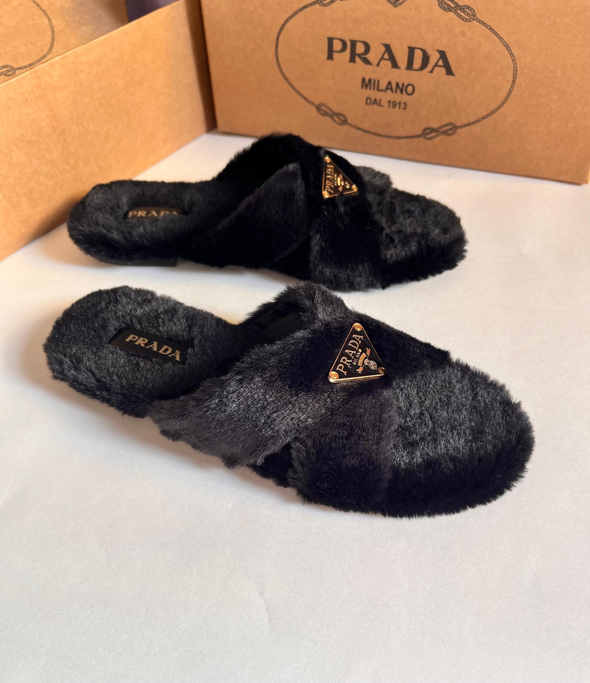 Black fuzzy slippers with Pra da logo on a white surface, with Pra da box in the background.