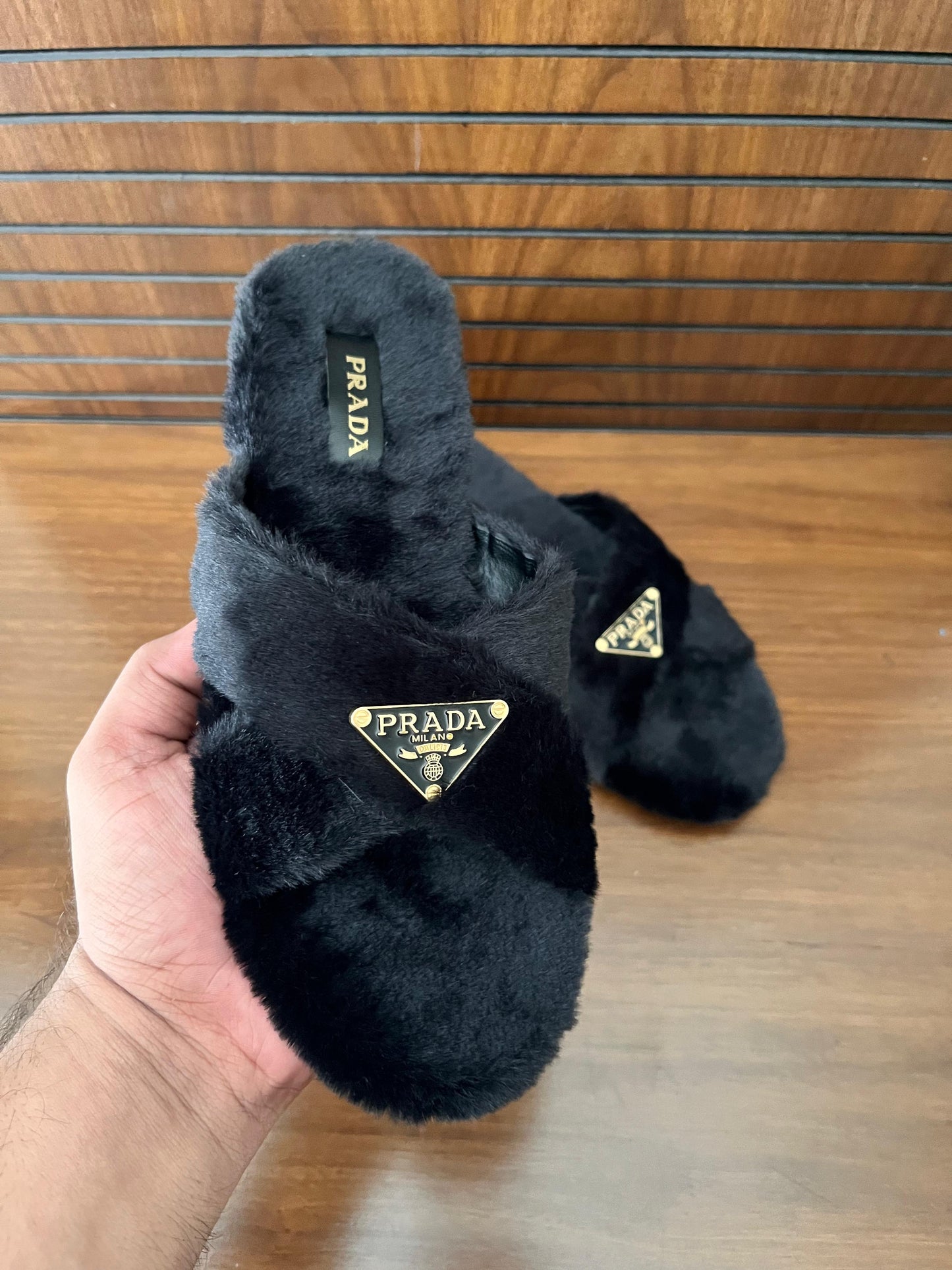 Black fuzzy Pra da slippers held by a hand on a wooden floor.