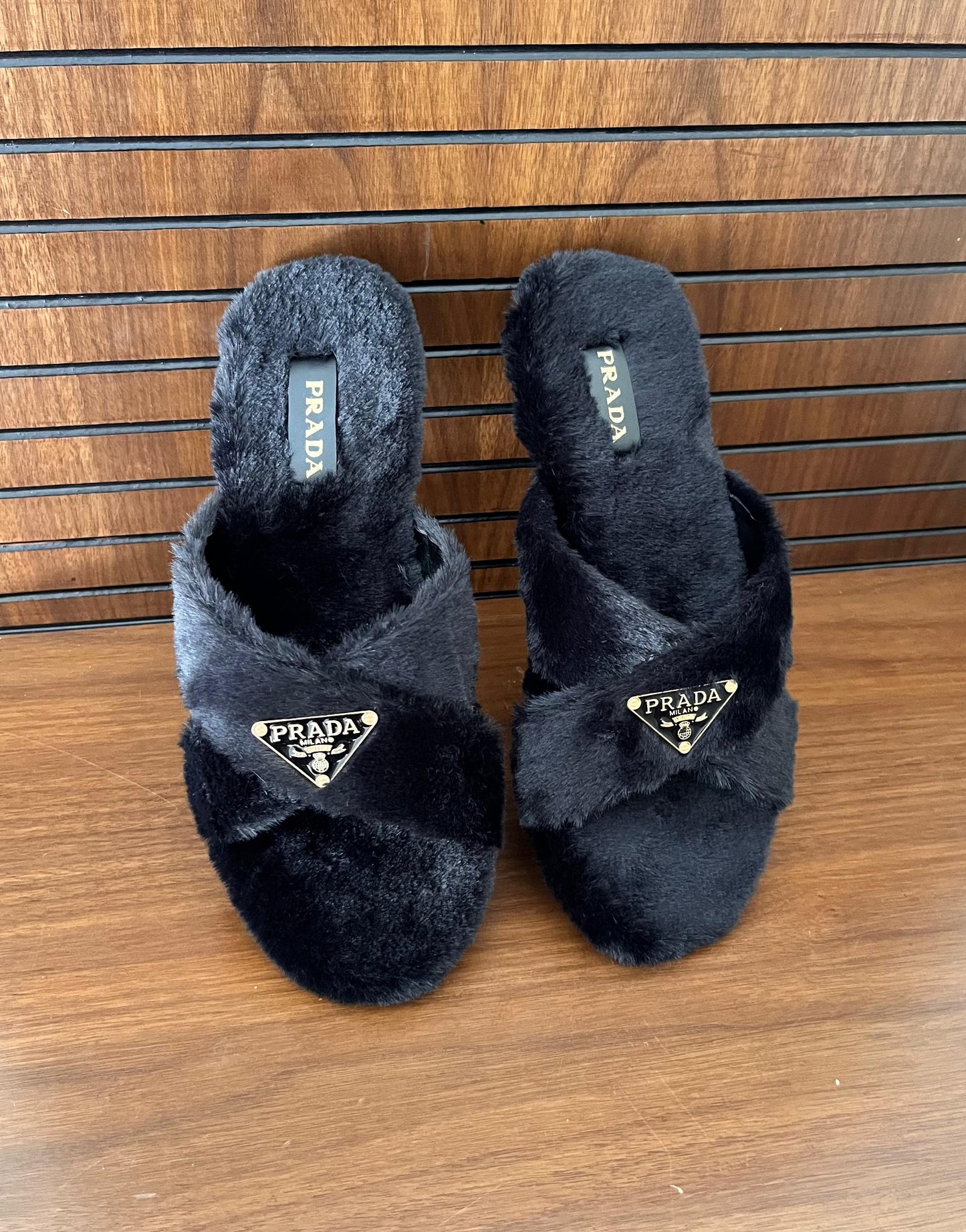 Pair of black fuzzy slippers with Pra da logo on a wooden floor.