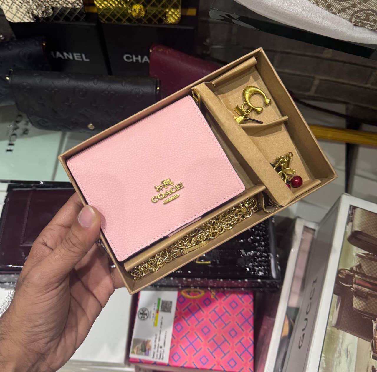Pink wallet with gold accents held in a hand, with a blurred background of fashion magazines and bags.