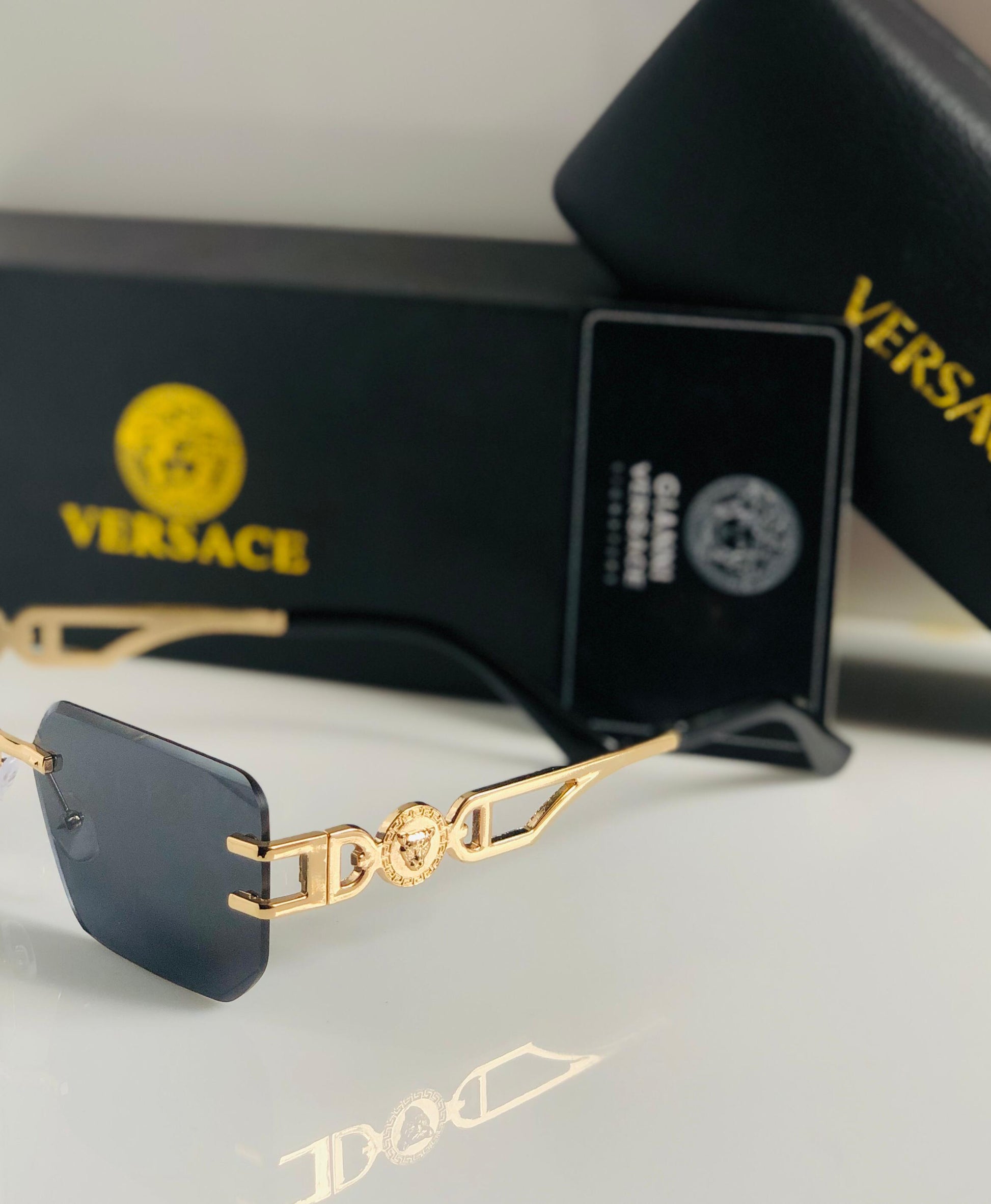 Ver sace sunglasses with gold frame and black lenses on a white surface, with Versace packaging in the background.