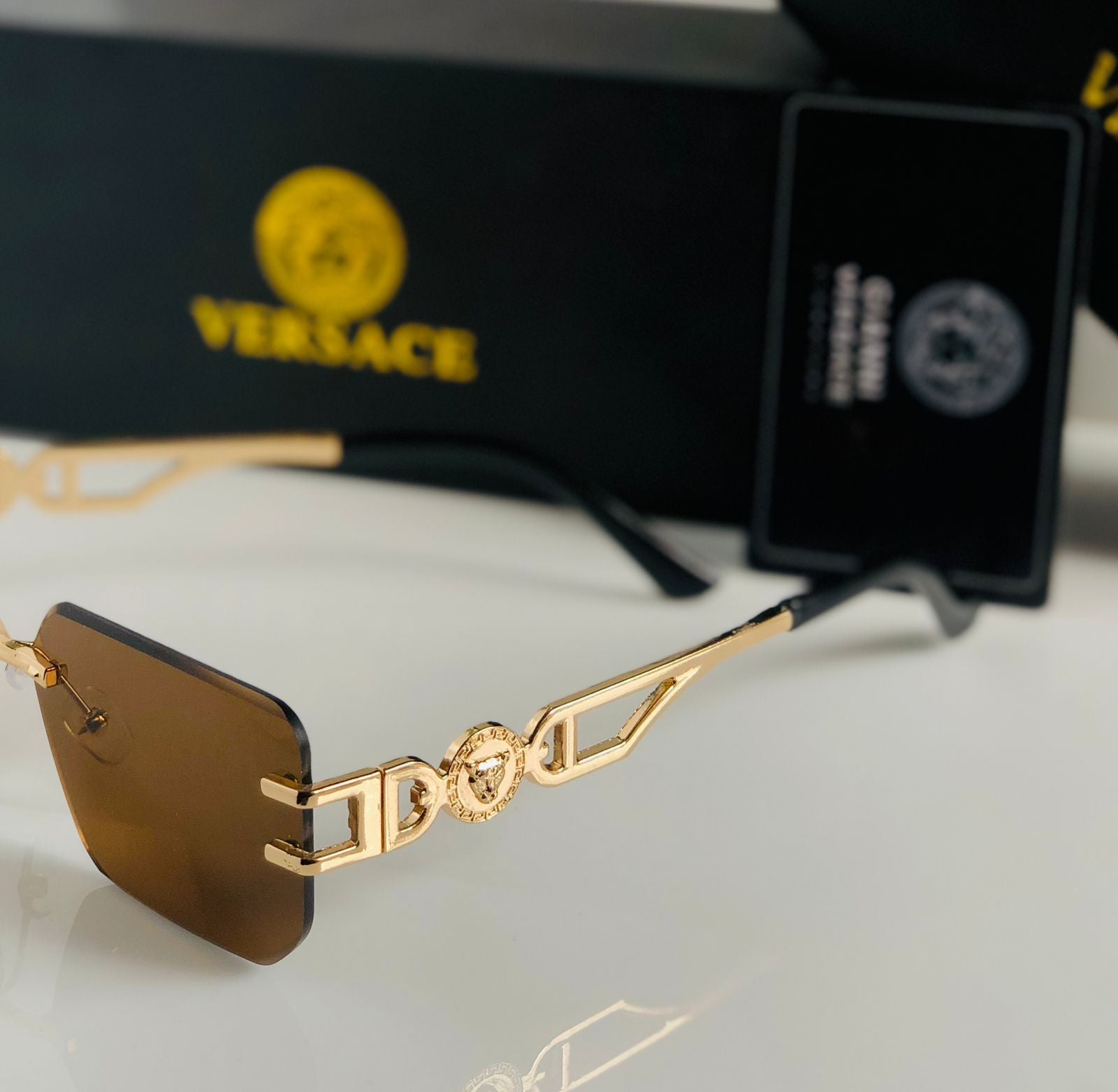 Gold-framed sunglasses with brown lenses on a white surface, Ver sace box in the background.