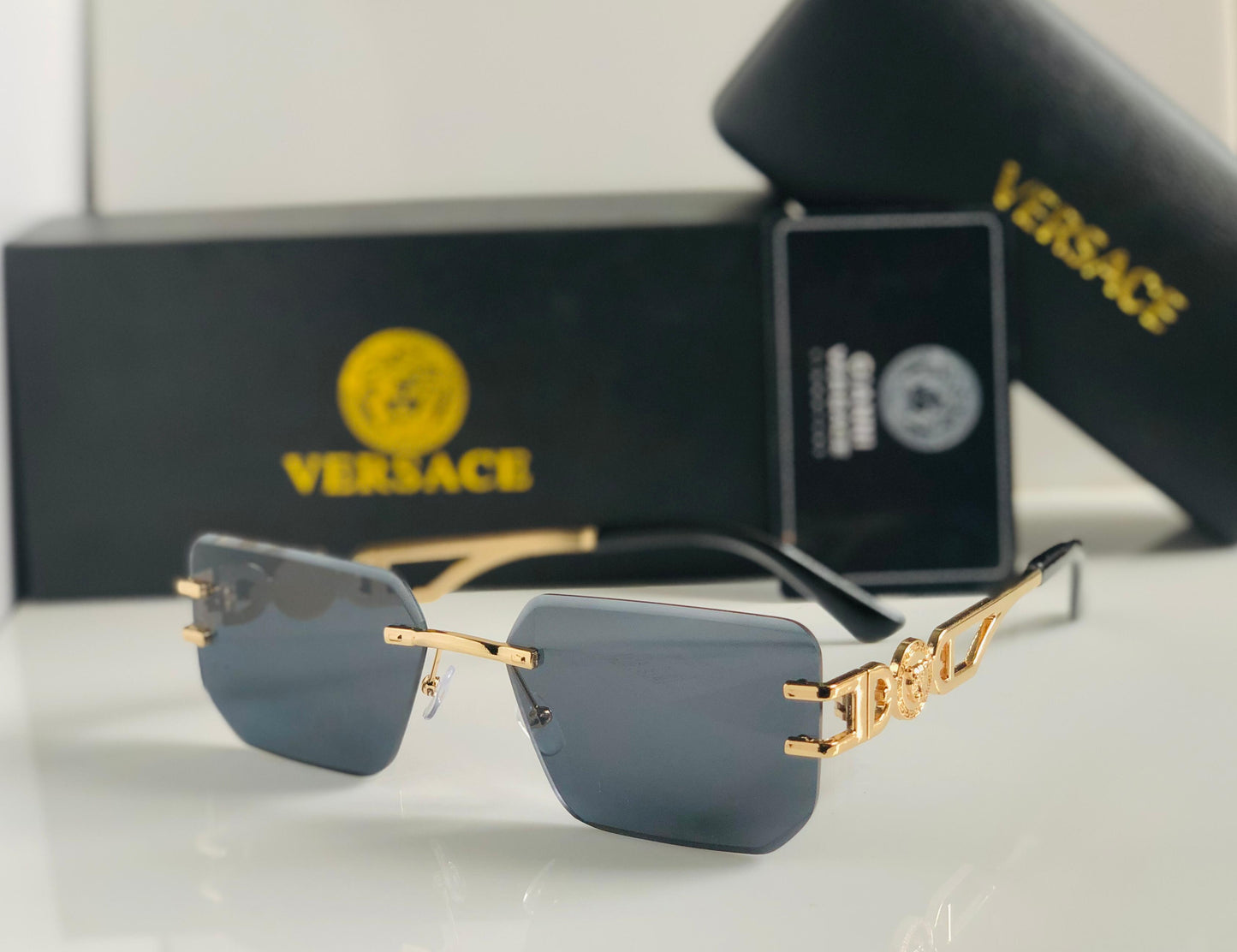 Ver sace sunglasses with gold frames and gray lenses on a white surface, with Versace packaging in the background.