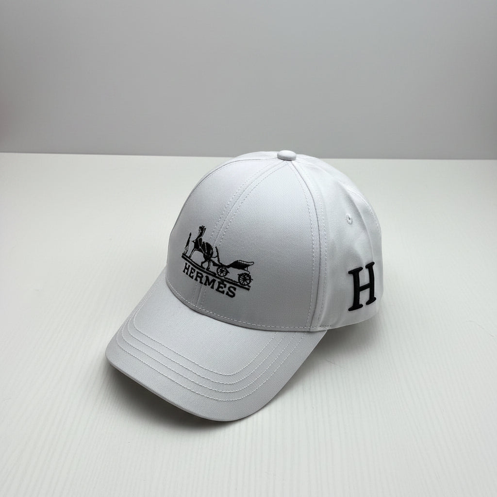 A white structured baseball cap with black embroidered Hermès logo and horse-and-carriage emblem on the front, plus a large black 'H' on the side, photographed on a white surface with an orange Hermès box in the background.