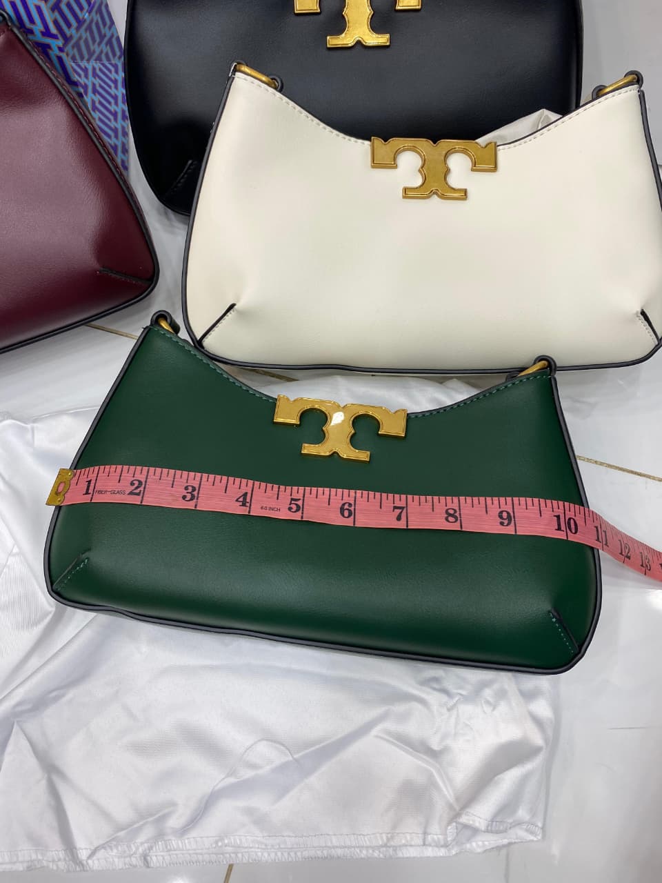 Three handbags in different colors with a gold logo, one being measured with a pink tape measure.
