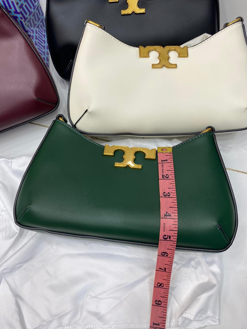 Green handbag with gold logo and white bag with gold logo, measured with a pink tape measure.