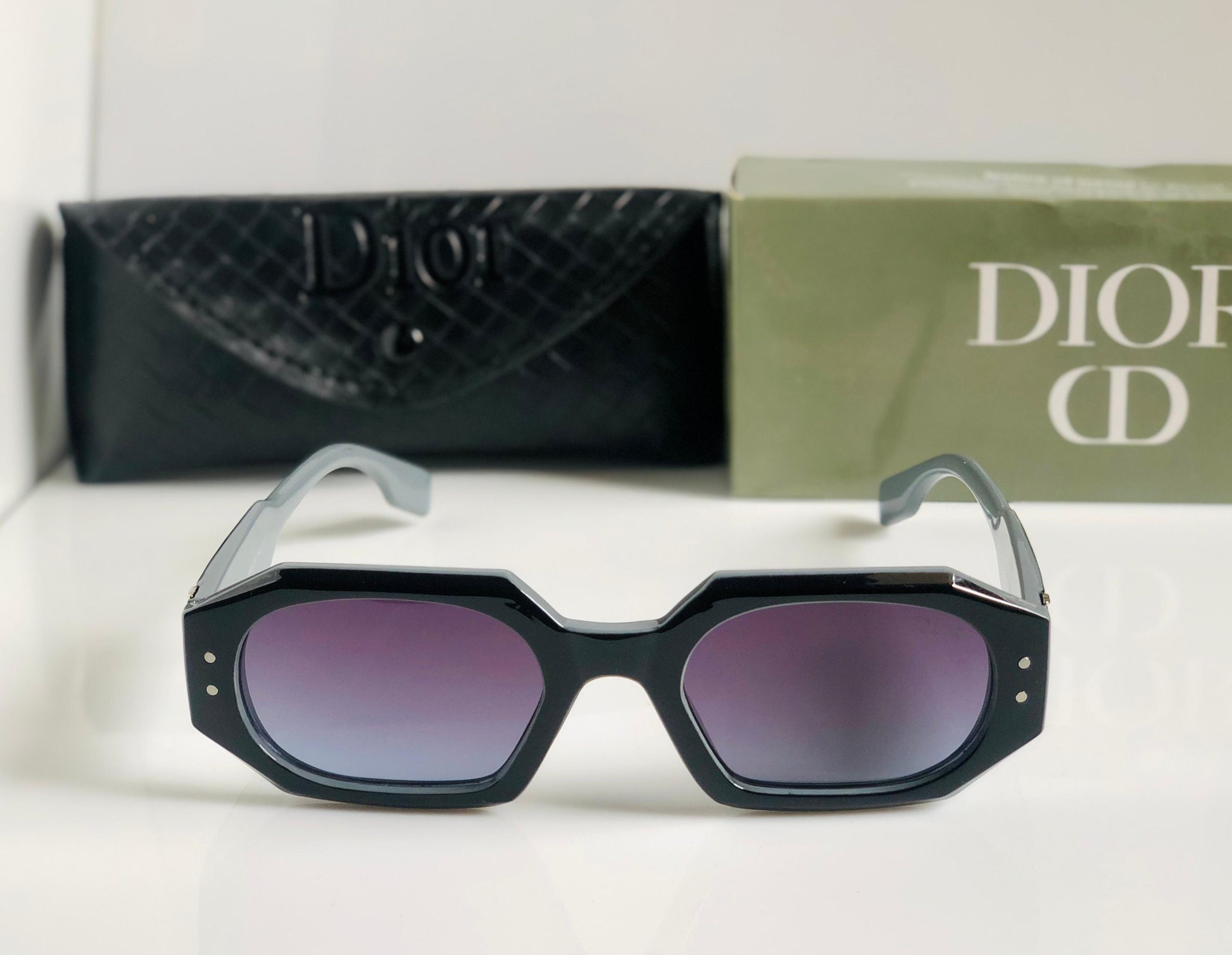 Black sunglasses with purple lenses in front of a Di or box and pouch.