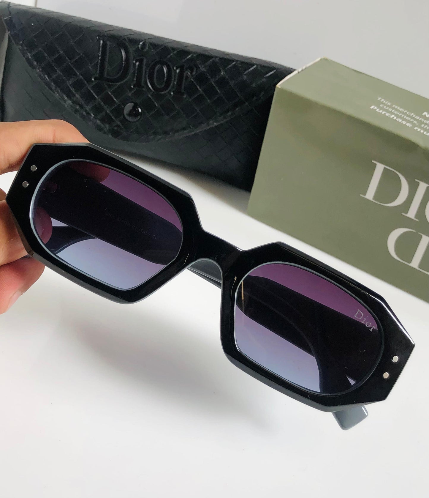 Black sunglasses held by a hand with Di or box and case in the background