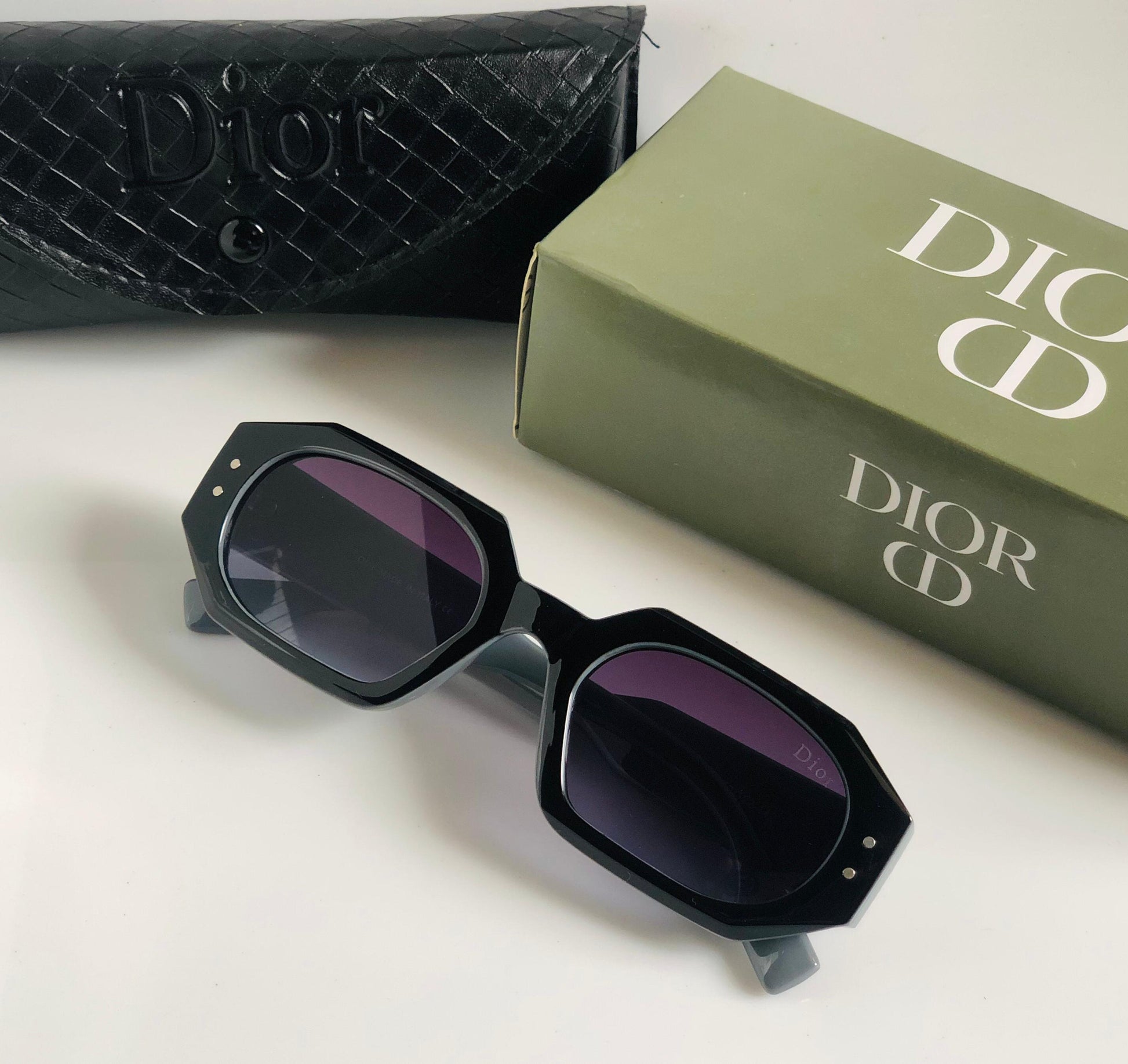Black sunglasses with purple lenses on a white surface next to a Di or box.