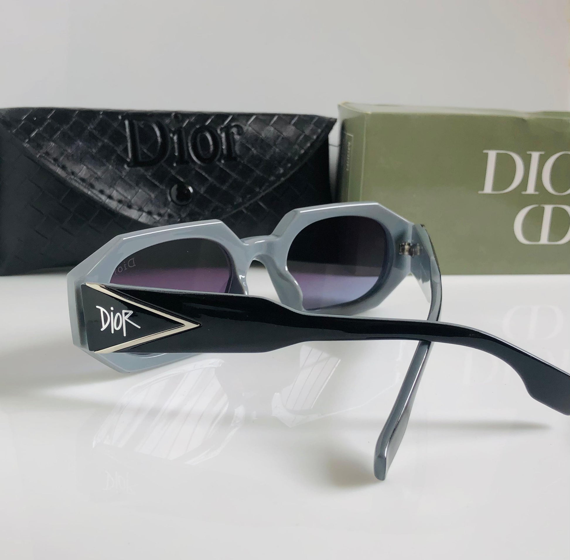 Di or sunglasses with black and silver frame on a white surface, with Di or bag and box in the background.