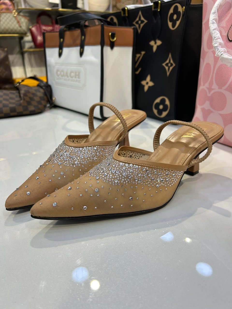 A pair of beige pointed-toe mule heels covered in silver rhinestones, displayed on a reflective white surface with Coach and Louis Vuitton shopping bags in the background.