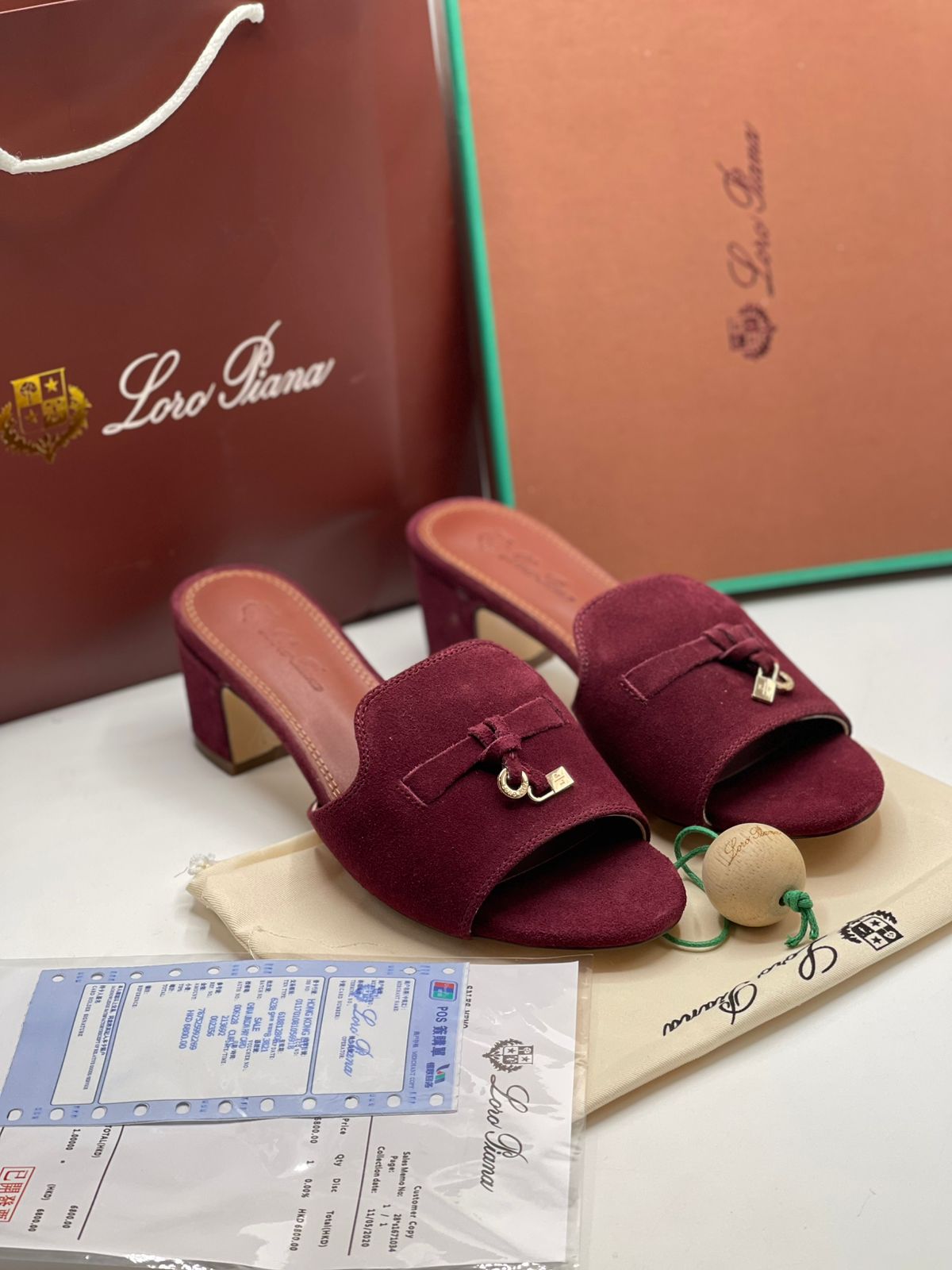 A pair of burgundy suede mule sandals with gold-tone hardware, displayed with their branded brown shopping bag, dust bag, and packaging box on a white surface.