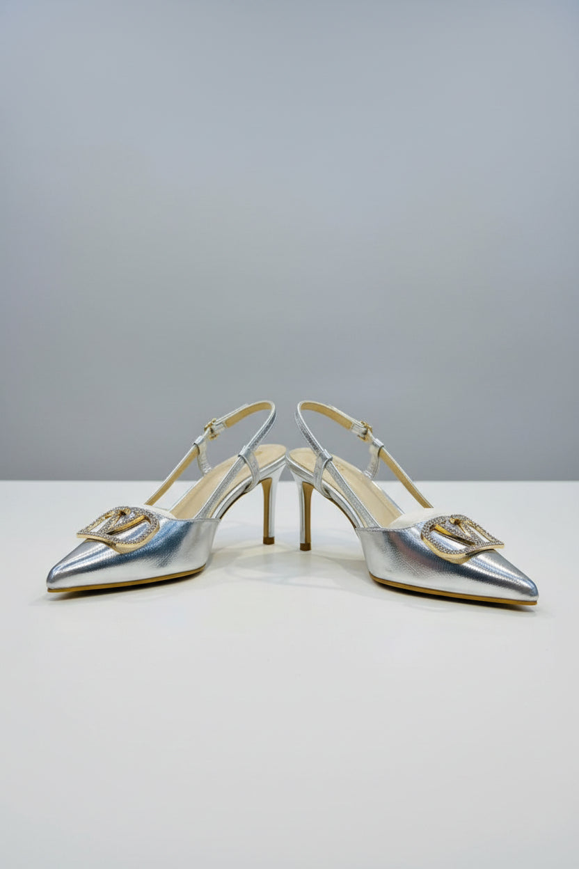 A pair of metallic silver pointed-toe slingback heels with large gold V-shaped hardware embellished with rhinestones, displayed on a white surface with a clear stand in a luxury handbag shop.