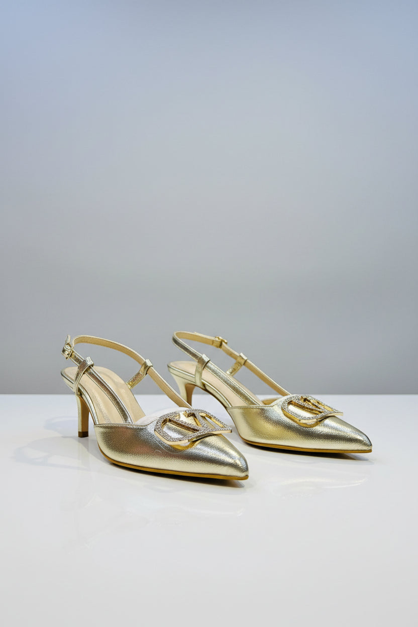 A pair of metallic gold pointed-toe slingback heels with large interlocking LV-style letter embellishments on the front, displayed on a white counter in a retail setting with shelves of handbags behind them.