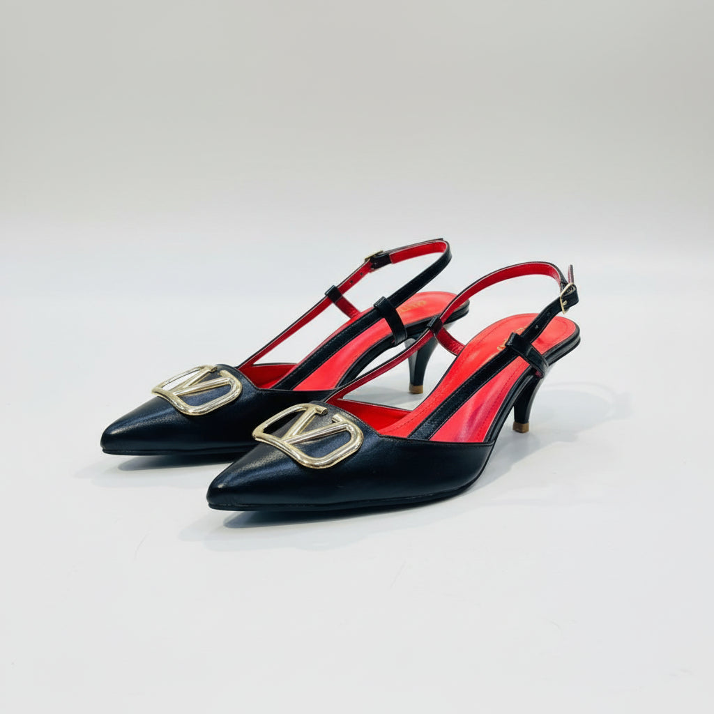 A pair of black leather pointed-toe slingback heels with a bright red insole and a large gold-tone metal buckle detail on the toe, displayed on a white counter in a shop with shelves of handbags in the background.