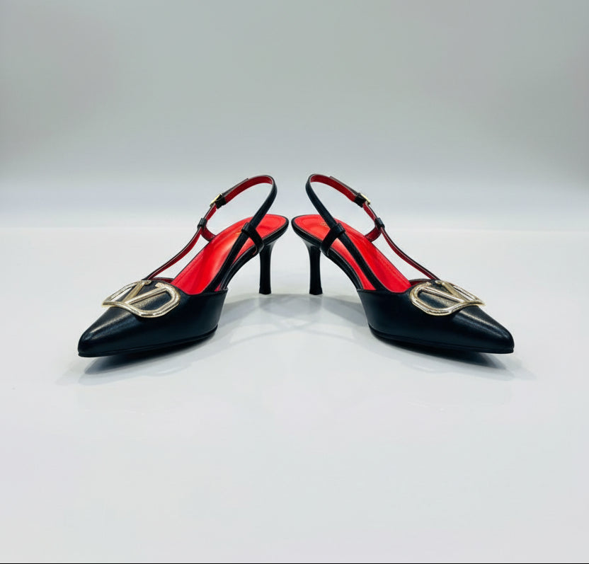 A pair of black leather slingback pointed-toe heels with a prominent gold-tone V logo on the toe and a bright red insole, displayed on a white counter with a blurred background of handbags.