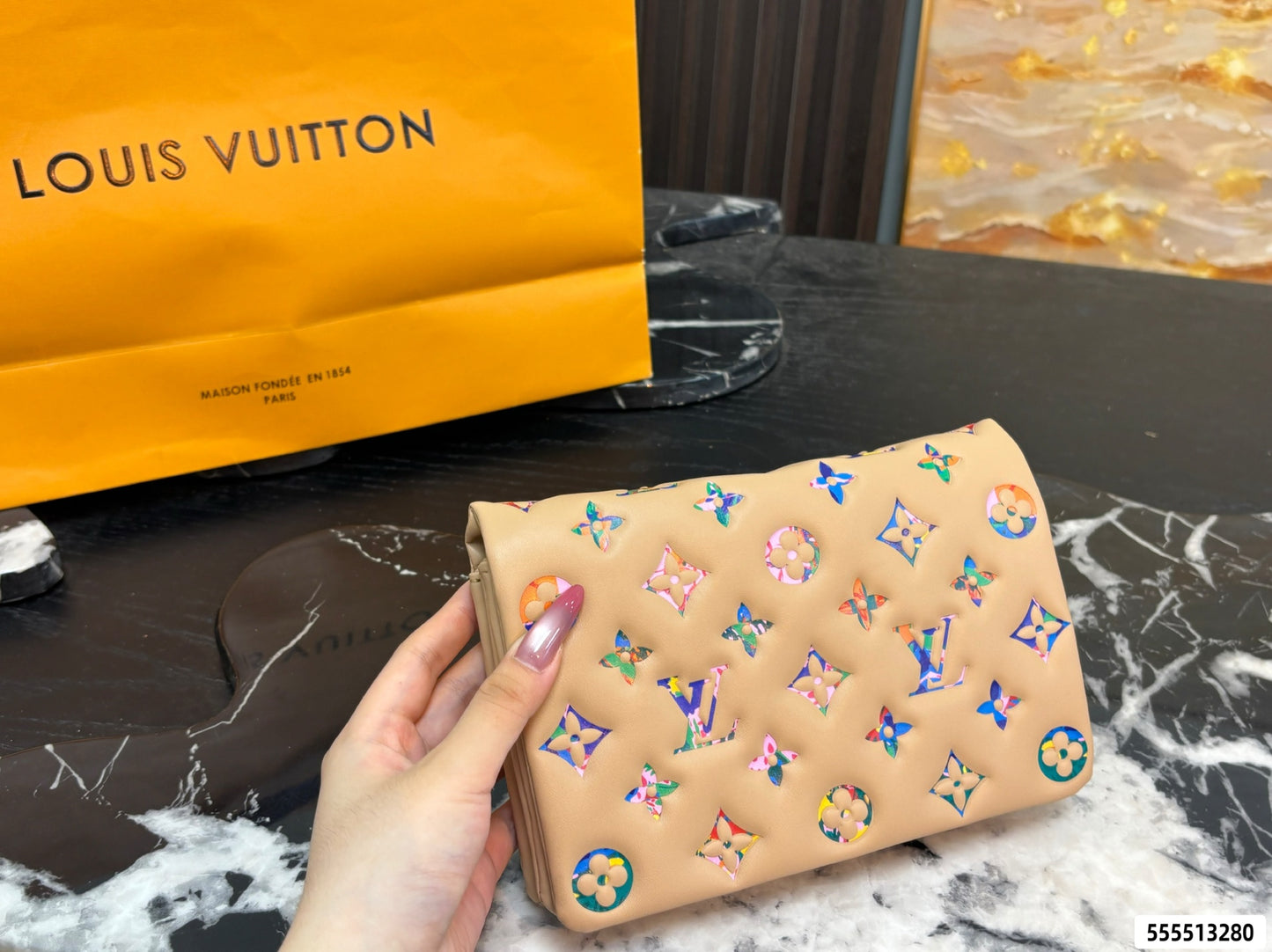 A hand holds a beige Louis Vuitton wallet decorated with colorful monogram patches, with a yellow Louis Vuitton shopping bag in the background on a black marble surface.