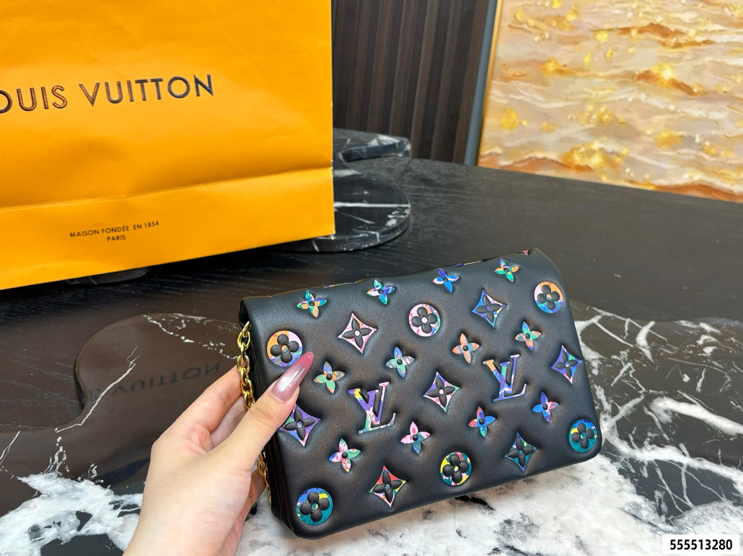 A hand holds a black Louis Vuitton shoulder bag covered in iridescent multicolor monogram patches. A yellow Louis Vuitton shopping bag and a brown leather pouch rest on a black marble surface in the background.
