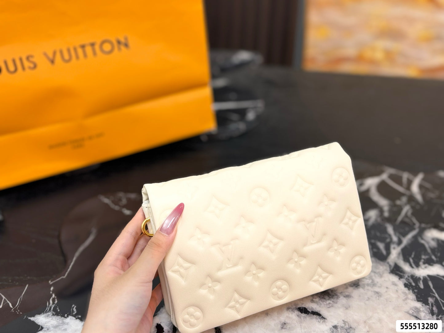 A hand holds a cream-colored Louis Vuitton monogram clutch bag, with a yellow Louis Vuitton shopping bag in the blurred background.